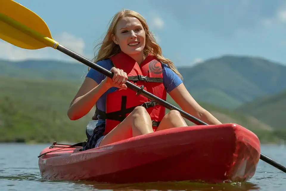 Woman kayaking on a lake, wearing a life jacket, with mountains in the background.
