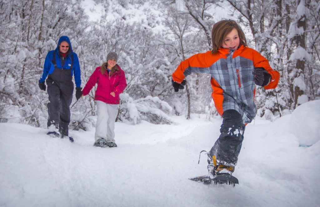 a kid running in snow shoes in the snow with two people holding hands in the background