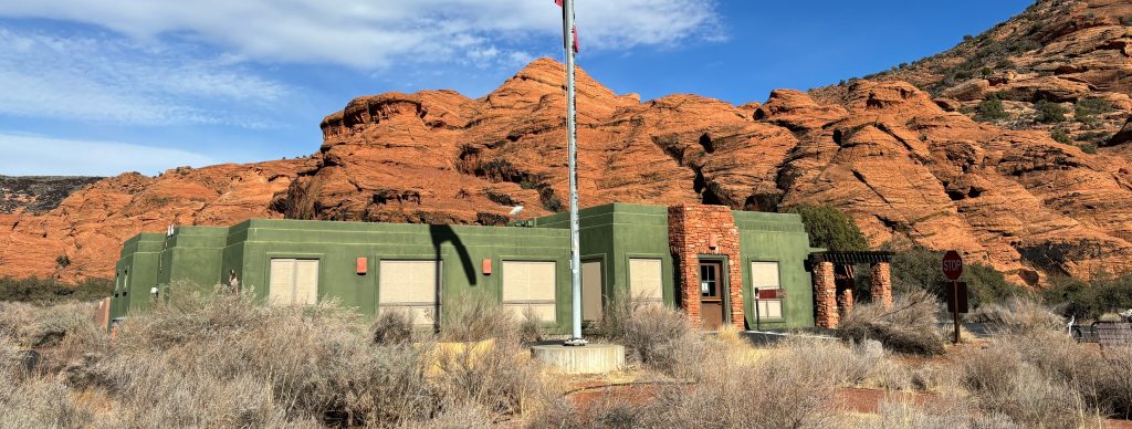 Picture of a green building with red rock cliffs behind it. There are brown bushes in the foreground.