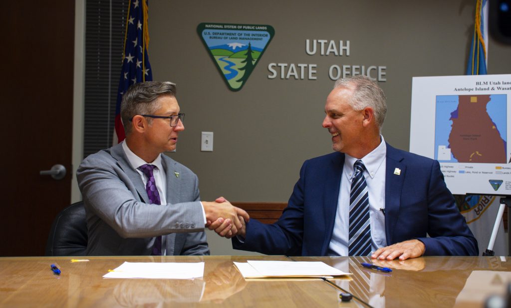 Two men in suits shaking hands with paper in front of them. In the background is an American flag, a BLM emblem, and a map of the land conveyance.